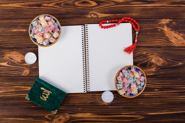 Free photo an overhead view of spiral notebook with delight lukum bowls; kuran and red prayer beads on wooden desk