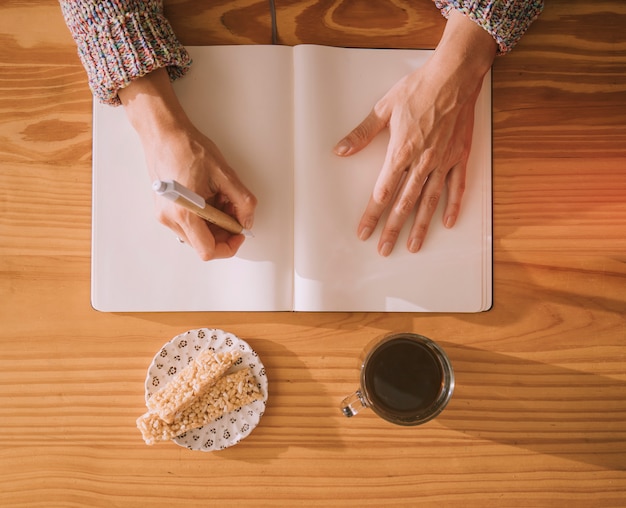 An overhead view of woman writing on blank white notebook with pen and breakfast on table