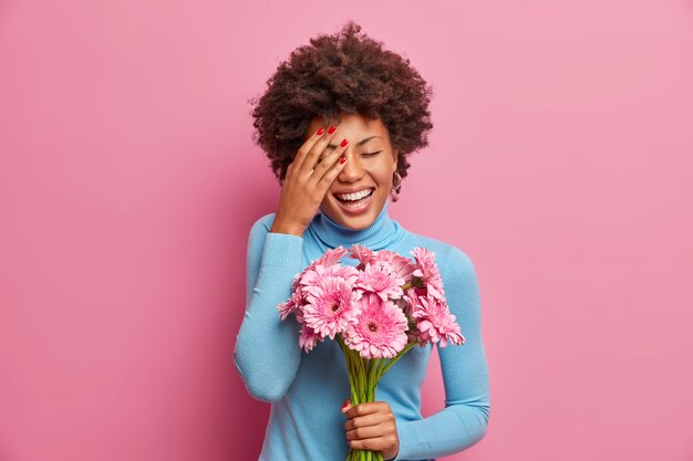 Overjoyed Afro American woman laughs out loudly, keeps palm on face, stands indoor with bouquet of gerbera flowers