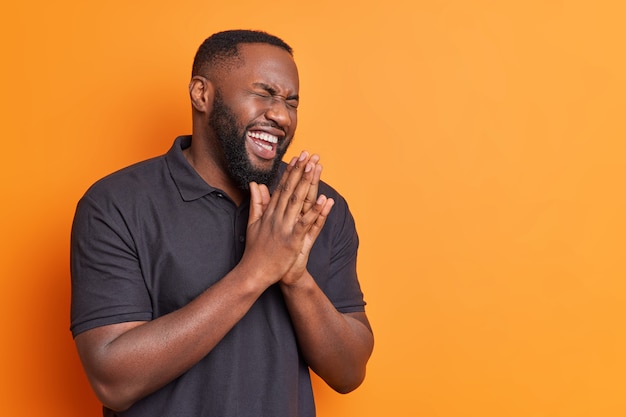 Overjoyed bearded adult man rubs palms and laughs out happily dressed in casual black t shirt hears funny joke poses against bright orange wall with copy space for your text