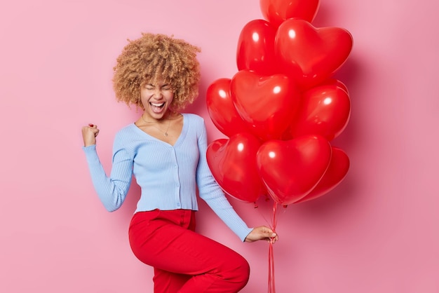 Free Photo overjoyed curly haired woman exclaims loudly clenches fist celebrates special occasion wears casual jumper and red trousers holds bunch of heart balloons poses against pink backgound happy holiday