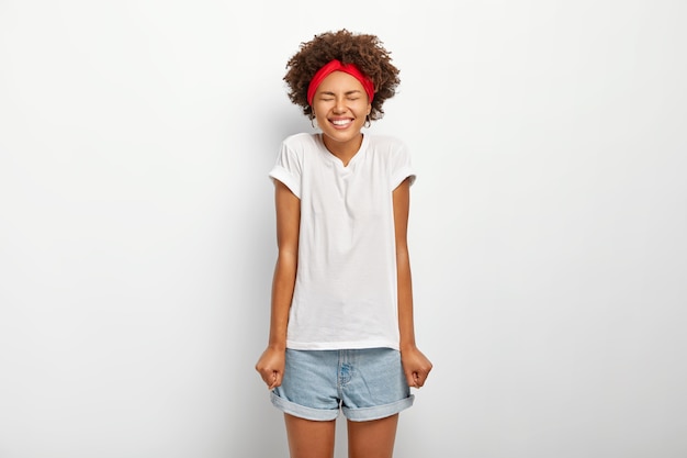 Overjoyed dark skinned woman with Afro hair keeps hands clenched in fists, enjoys long awaited holidays, dressed in casual summer outfit, expresses good emotions, isolated on white background