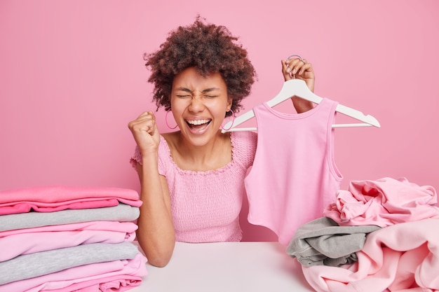 Overjoyed dark skinned woman with curly hair cleches fist from happiness holds apparel on hanger sits at table folds laundry isolated over pink wall