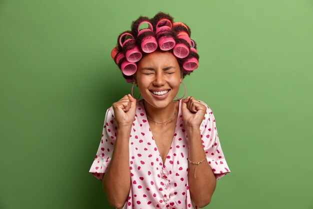 Overjoyed dark skinned young woman clenches fists, closes eyes and smiles broadly, rejoices good news, spends free time at home, wear hair rollers, makes hairstyle, isolated on green 