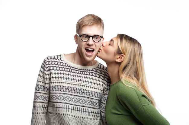 Overjoyed emotional young bearded male nerd wearing eyeglasses exclaiming excitedly, being shocked while beautiful woman kissing him on cheek