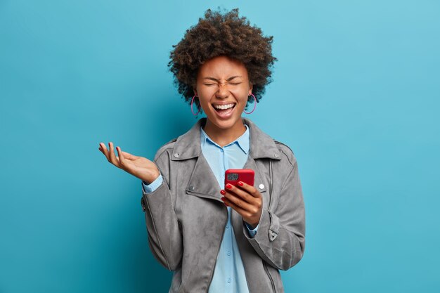 Overjoyed ethnic woman with curly hair, laughs as watches funny video on smartphone, closes eyes and raises hand, being in good mood, wears grey casual jacket, poses 