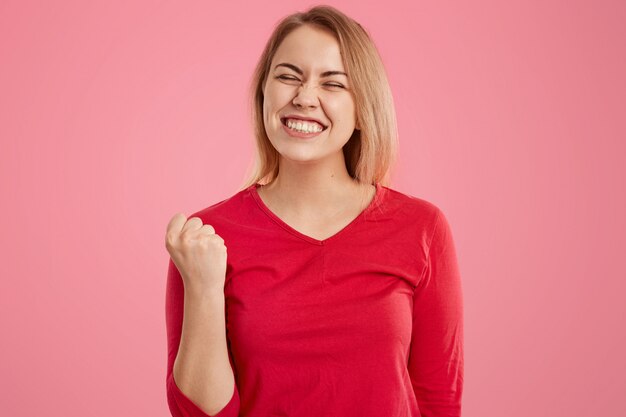 Overjoyed European woman with short hair, clenches fist, dressed in red casual outfit, squints face, dressed in red jumper