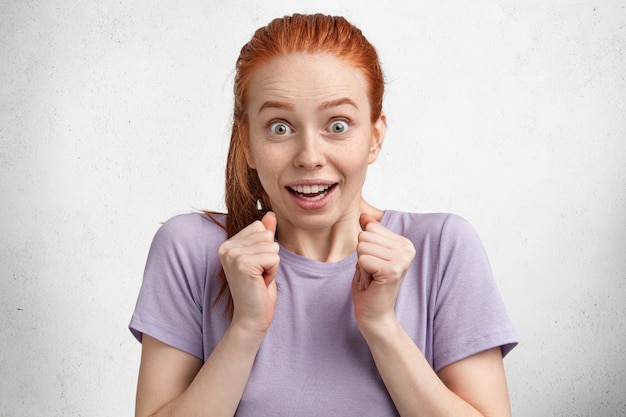 Free photo overjoyed excited ginger young female model in purple casual t shirt, gestures actively, keeps hands in fists, demonstrates surprised reaction on what she hears