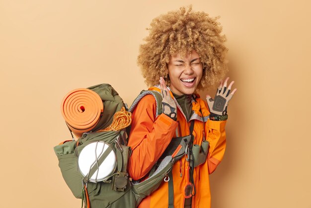 Overjoyed female tourist exclaims loudly keeps hands raised wears gloves orange jacket carries heavy rucksack with necessary equipment isolated over brown background Summer time adventure concept