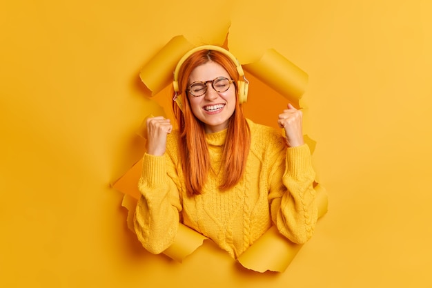 Free photo overjoyed ginger woman closes eyes and makes yes gesture listens favorite music via headphones celebrates successful deal feels relaxed amused.