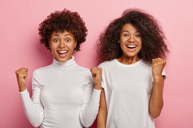 Overjoyed happy two stundents with Afro hair, keeps arms raised, clench fists with success