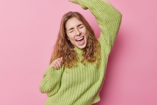 Free photo overjoyed long haired woman dances carefree exclaims from joy moves with rhythm of music wears loose knitted sweater isolated over pink background catches nice moment has fun enjoys freedom.