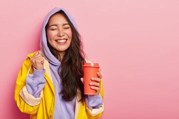 Overjoyed pleased Asian woman raises clenched fist, keeps eyes closed, drinks hot beverage from flask, rejoices happy moment, wears warm waterproof clothes