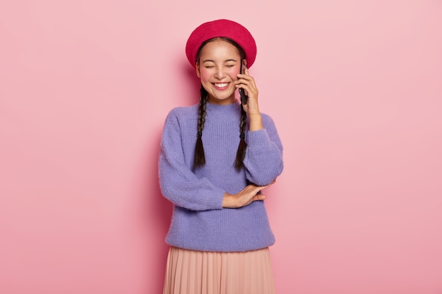 Overjoyed pleased woman with Asian appearance, enjoys funny telephone conversation with friend, keeps modern cellular near ear, has two long pigtails