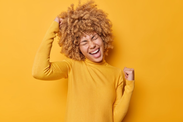 Free photo overjoyed positive curly haired woman makes yes gesture cheers and celebrates success wears scasual jumper feels like winner isolated over vivid yellow background people and triumph concept