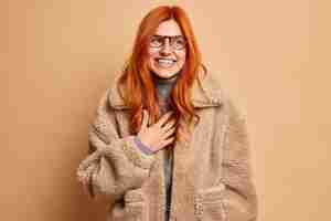 Free photo overjoyed redhead adult woman laughs and expresses happy sincere emotions wears glasses and warm brown fur coat concentrated aside with smile enjoys winter time has upbeat mood. fashion concept