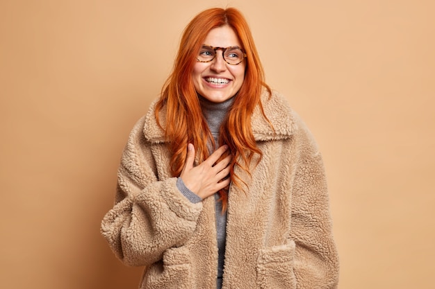 Free Photo overjoyed redhead adult woman laughs and expresses happy sincere emotions wears glasses and warm brown fur coat concentrated aside with smile enjoys winter time has upbeat mood. fashion concept