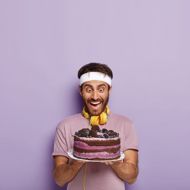 Free photo overjoyed surprised man stares at delicious cake, feels temptation, being hungry after active workout, wears casual t shirt, carries headphones