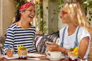 Free photo overjoyed two women laugh happily while share opininos about planning project, communicate during coffee break, write records in organizer, eat delicious dessert, wear casual clothes and spectacles