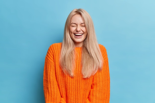 Free photo overjoyed woman with blonde long hair laughs positively hears something funny closes eyes shows white teeth wears orange knitted sweater