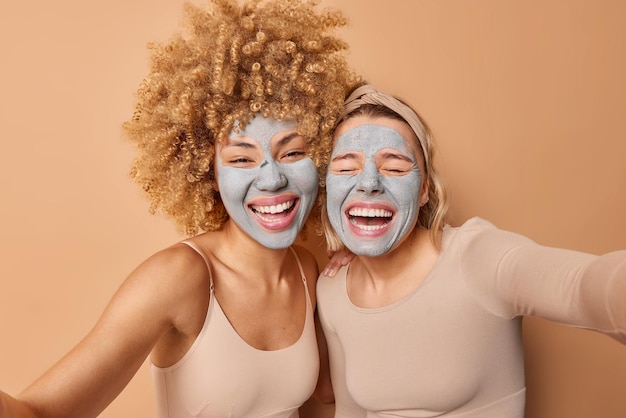 Free Photo overjoyed women laugh gladfully pose next to each other have fun make selfie photo keep arms outstreched take care of skin wears nourishing clay beauty masks isolated over brown wall beauty concept