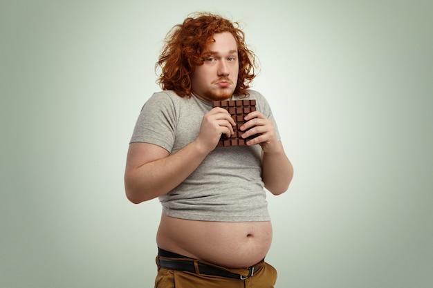 Free photo overweight fat man with ginger curly hair looking indecisive and hesitant, holding large bar of chocolate with both hands while forbidden to eat sugar, and junk food because of strict low-carb diet