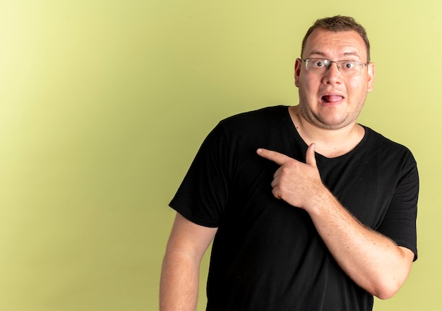 Overweight man in glasses wearing black t-shirt happy and surprised pointing with finger to the side standing over light wall