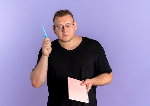 Overweight man in glasses wearing black t-shirt holding notebook and pen looking puzzled standing over blue wall