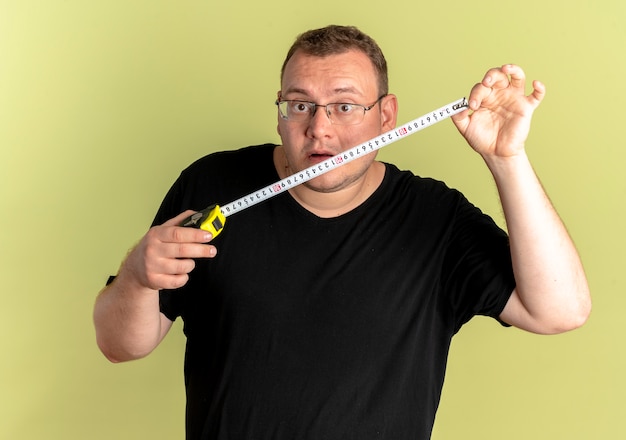 Overweight man in glasses wearing black t-shirt holding ruler looking at it surprised over light