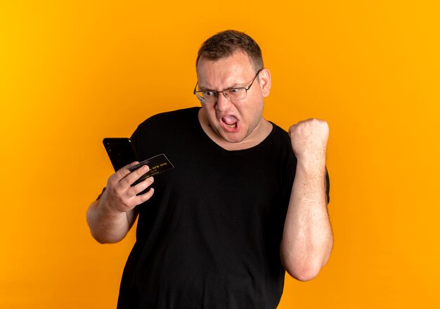 Overweight man in glasses wearing black t-shirt holding smartphone clenching fist shouting over orange