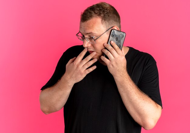 Overweight man in glasses wearing black t-shirt looking aside confused while talking on mobile phone over pink