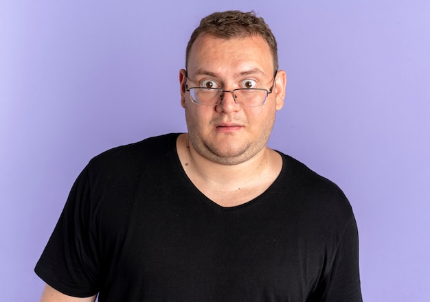 Overweight man in glasses wearing black t-shirt looking at camera surprised standing over blue wall