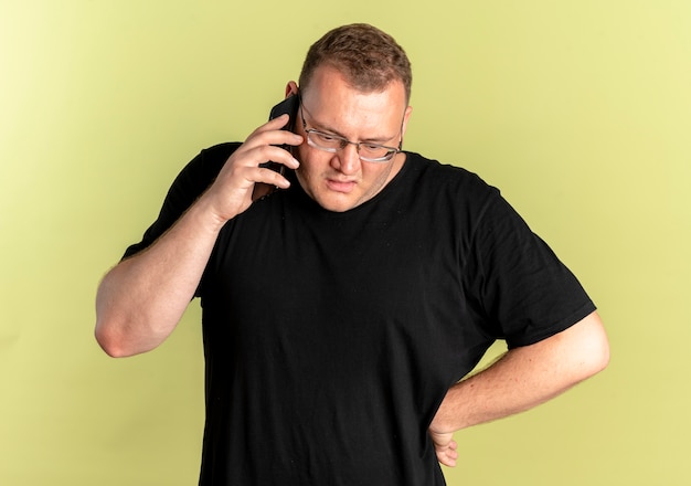 Free Photo overweight man in glasses wearing black t-shirt looking confused and displeased while talking on mobile phone standing over light wall