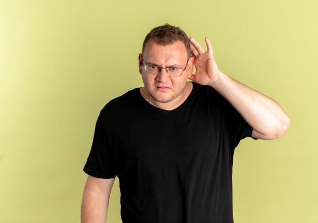 Free Photo overweight man in glasses wearing black t-shirt looking confused holding his palm near ear trying to listen standing over light wall