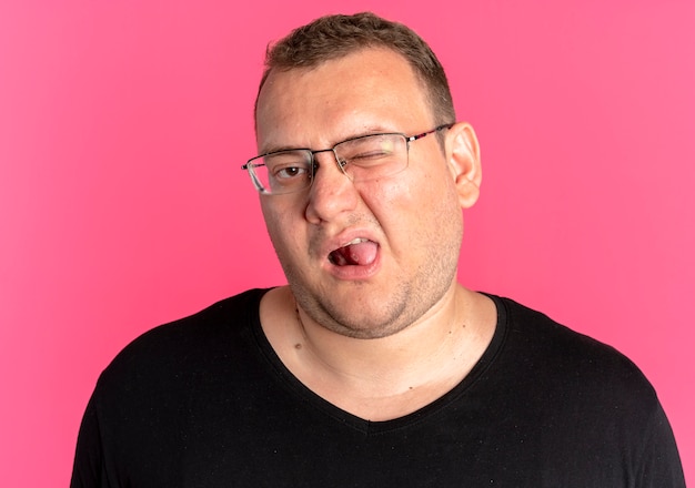 Overweight man in glasses wearing black t-shirt looking displeased sticking out tongue over pink