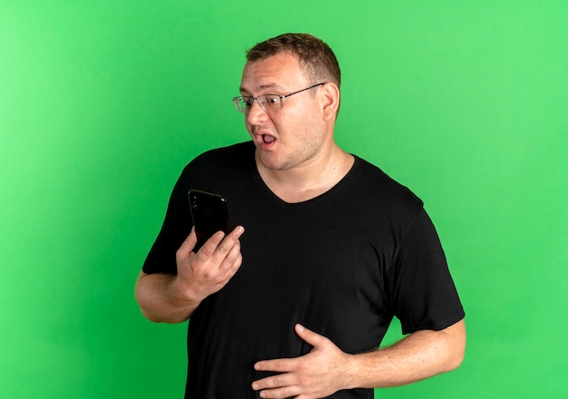 Free photo overweight man in glasses wearing black t-shirt looking at his smartphone screen confused and surprised over green