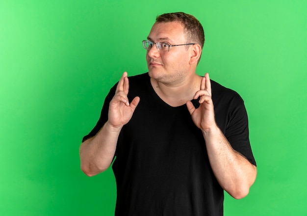 Free photo overweight man in glasses wearing black t-shirt making desirable wish crossing fingers with happy face over green