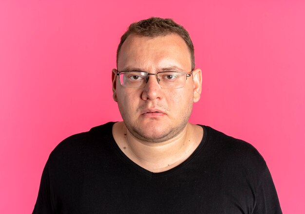 Overweight man in glasses wearing black t-shirt with serious face over pink