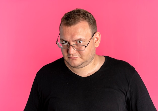 Overweight man in glasses wearing black t-shirt  with serious face standing over pink wall