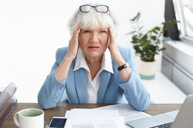 Overwhelmed frustrated mature elderly European female accountant wearing formal suit having painful stressed look because of mistake in financial report, massaging temples, suffering from headache