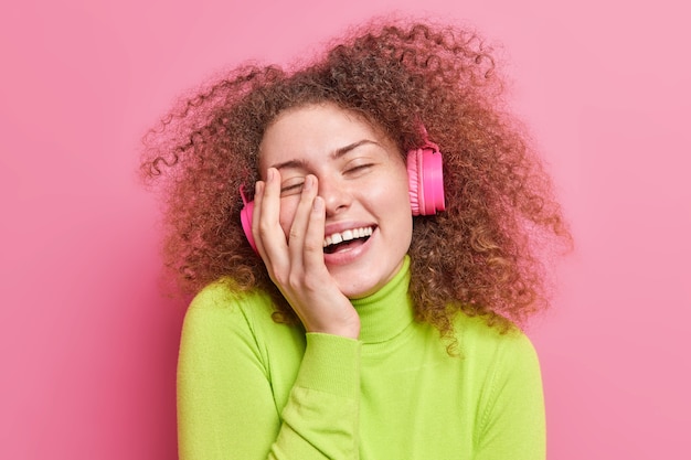 Free Photo overyoyed teenage girl with curly hbushy hair smiles broadly keeps hand on face closes eyes from pleasure enjoys listening music via wireless headphones dressed casually isolated on pink wall