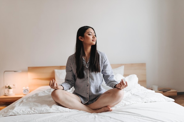 Pacified longhaired girl in pajamas is meditating Snapshot of Asian in bright bedroom