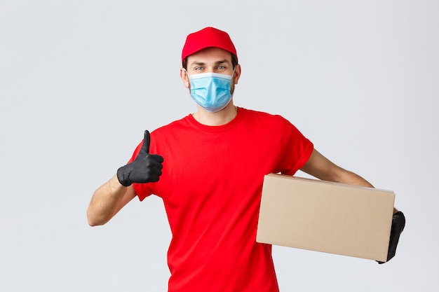 Packages and parcels delivery, covid-19 quarantine delivery, transfer orders. Cheerful courier in red uniform, gloves and face mask, thumb-up, recommend contactless deliver, holding box with order