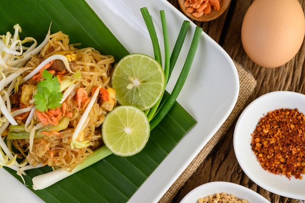 Pad Thai in a white plate with lemon, eggs and seasoning on a wooden table.