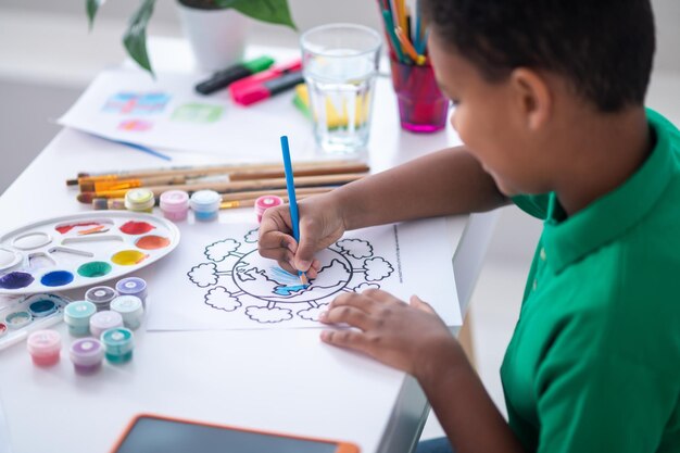 Painting. Dark-skinned primary school-age boy drawing with blue pencil sideways to camera while sitting at table in room under daylight