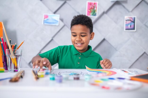 Paints. Smiling dark-skinned school-age boy in casual green t-shirt sitting at table with art supplies touching tube of paint in light room