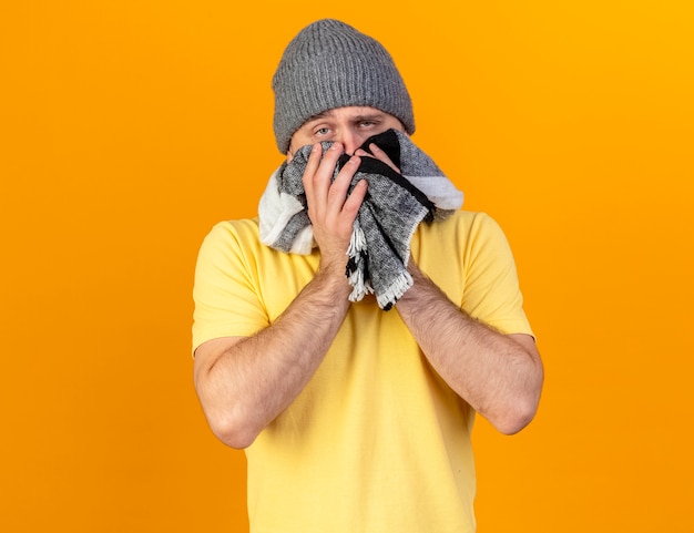 Free photo pale young blonde ill slavic man wearing winter hat and holds scarf isolated on orange wall with copy space