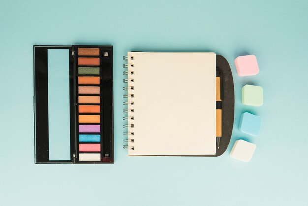 Palette of make up with colorful sponge and blank spiral notepad on colored backdrop