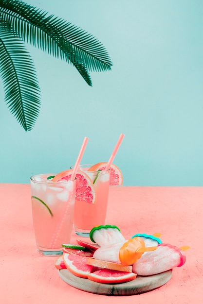 Free photo palm leaves over the grapefruit cocktail glasses; popsicles on coral desk against teal background