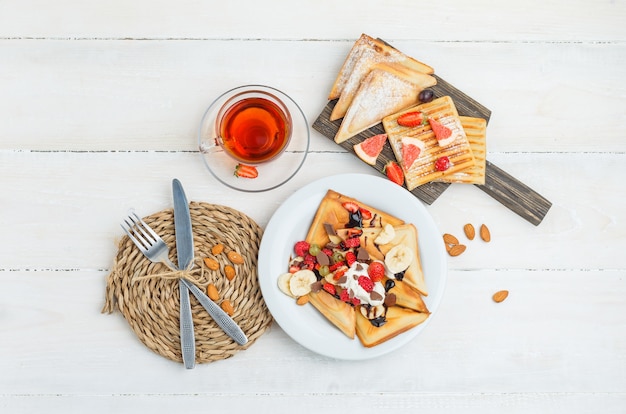 Pancakes on wooden board with tea, almonds, knife, fork, grapes and raspberries